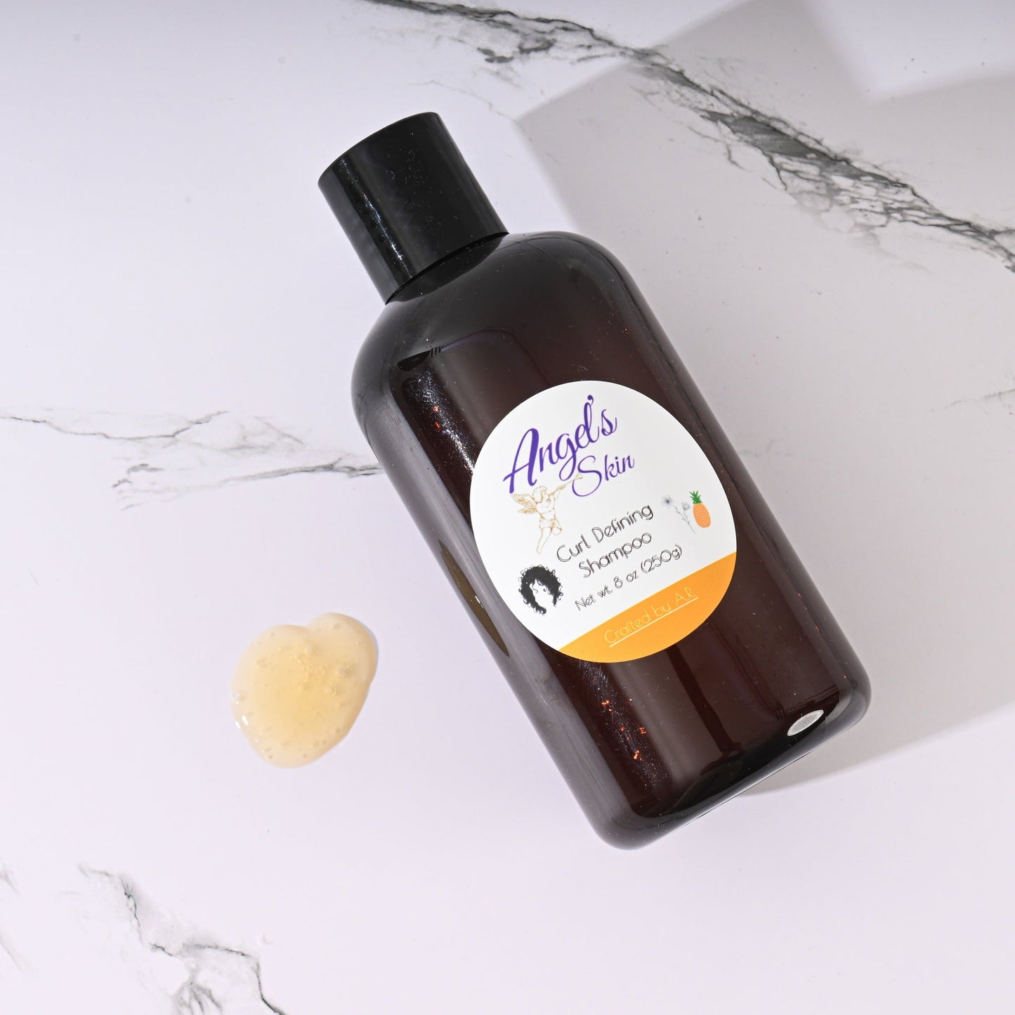 Bottle of Angel's Skin Curl Defining Shampoo with pineapple and flaxseed on marble surface with shampoo drop nearby